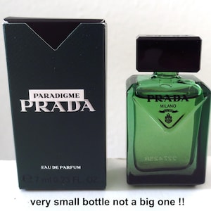 Könnte beinhalten: Ein grünes Prada Parfümfläschchen und die passende Schachtel. Die Flasche hat einen schwarzen Deckel und die Schachtel trägt die weißen Worte "PARADIGME PRADA". Die Flasche enthält 7 ml.