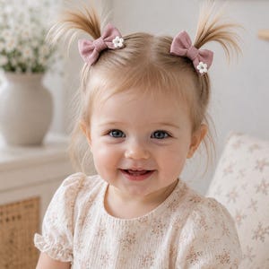 May include: A smiling toddler with blonde hair styled in two pigtails, each secured with a mauve bow and a small white flower accent. The child wears a cream-colored dress with a floral pattern.