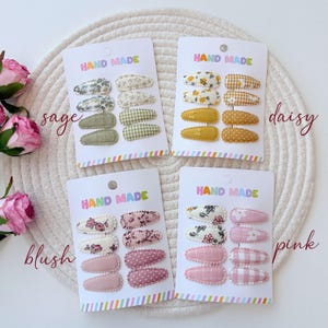 Op de afbeelding: Vier sets haarspeldjes op witte presentatiekaarten met het opschrift HAND MADE. Elke kaart bevat acht met stof beklede klikspeldjes in salie, madeliefje, blush en roze kleurenpaletten, met bloemen-, ruitjes-, stippen- en effen patronen.