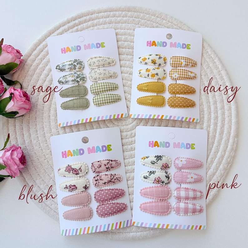 Op de afbeelding: Vier sets haarspeldjes op witte presentatiekaarten met het opschrift HAND MADE. Elke kaart bevat acht met stof beklede klikspeldjes in salie, madeliefje, blush en roze kleurenpaletten, met bloemen-, ruitjes-, stippen- en effen patronen.