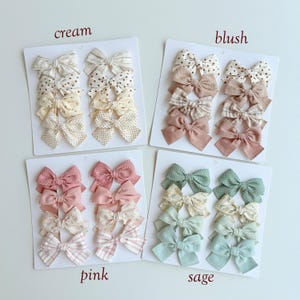 Peut inclure: Quatre cartes de pr&eacute;sentation blanches avec des n&oelig;uds pour cheveux en tissu dans des tons cr&egrave;me, blush, rose et sauge. Chaque carte contient huit n&oelig;uds aux motifs vari&eacute;s, incluant des pois, des rayures et des unis, avec les textes cream, blush, pink, sage.