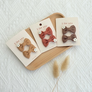 May include: Three sets of handmade hair bows in shades of brown and rust, each adorned with a small white flower. The bows are displayed on cream-colored cards that read "I like you" and "Hand Made". The bows are arranged on a wooden tray.
