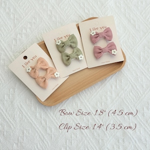 May include: Three sets of fabric hair bows in peach, sage green, and mauve, each adorned with a small white flower. The bows are displayed on cards that read "I like you". Bow size is 1.8 inches (4.5 cm).