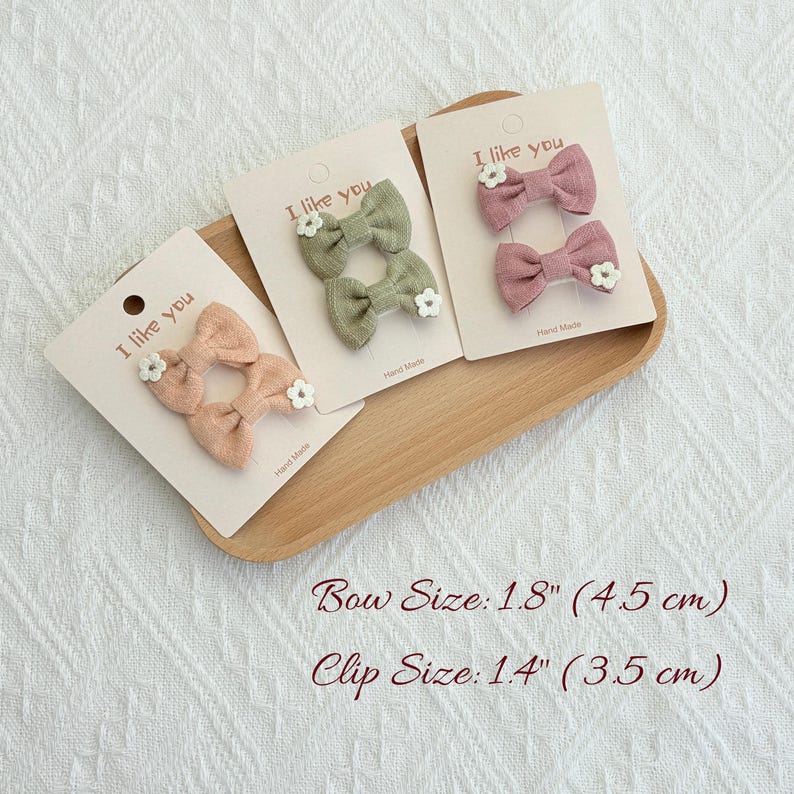 May include: Three sets of fabric hair bows in peach, sage green, and mauve, each adorned with a small white flower. The bows are displayed on cards that read "I like you". Bow size is 1.8 inches (4.5 cm).