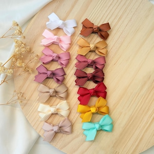 May include: A collection of small hair bows made from ribbed grosgrain ribbon, arranged in two vertical columns. The bows feature a variety of solid colors including white, pink, mauve, tan, cream, brown, mustard, maroon, red, yellow, and teal.