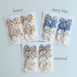 Peut inclure: Trois cartes de pr&eacute;sentation blanches avec des ensembles de n&oelig;uds pour cheveux en tissu. L'ensemble honey pr&eacute;sente des motifs floraux jaunes, &agrave; pois et vichy. L'ensemble dusty blue inclut des motifs floraux bleus, &agrave; pois et &agrave; carreaux. L'ensemble oatmeal montre des motifs &agrave; carreaux et &agrave; pois neutres.