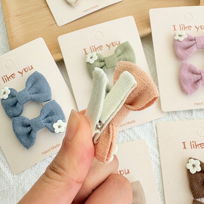 May include: A collection of handmade hair clips in various pastel colors, including blue, pink, and peach. Each clip features a bow and a small white flower. The cards behind the clips say "I like you".