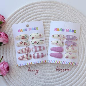 Op de afbeelding: Twee witte presentatiekaarten met het opschrift HAND MADE, elk met acht met stof beklede klikhaarspeldjes. De linkerkaart, gelabeld berry, bevat rood-witte patronen met kersen, bloemen en ruitjes. De rechterkaart, gelabeld blossom, toont bloemenprints en paarse ruitjes.