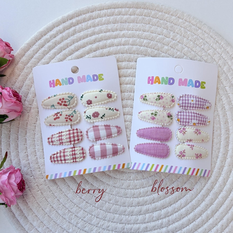 Op de afbeelding: Twee witte presentatiekaarten met het opschrift HAND MADE, elk met acht met stof beklede klikhaarspeldjes. De linkerkaart, gelabeld berry, bevat rood-witte patronen met kersen, bloemen en ruitjes. De rechterkaart, gelabeld blossom, toont bloemenprints en paarse ruitjes.