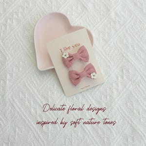 May include: Two pink bow hair clips with white floral accents are displayed on a light pink card that reads "I like you". The card is placed on a heart-shaped dish. The text "Delicate floral designs inspired by soft nature tones" is also visible.