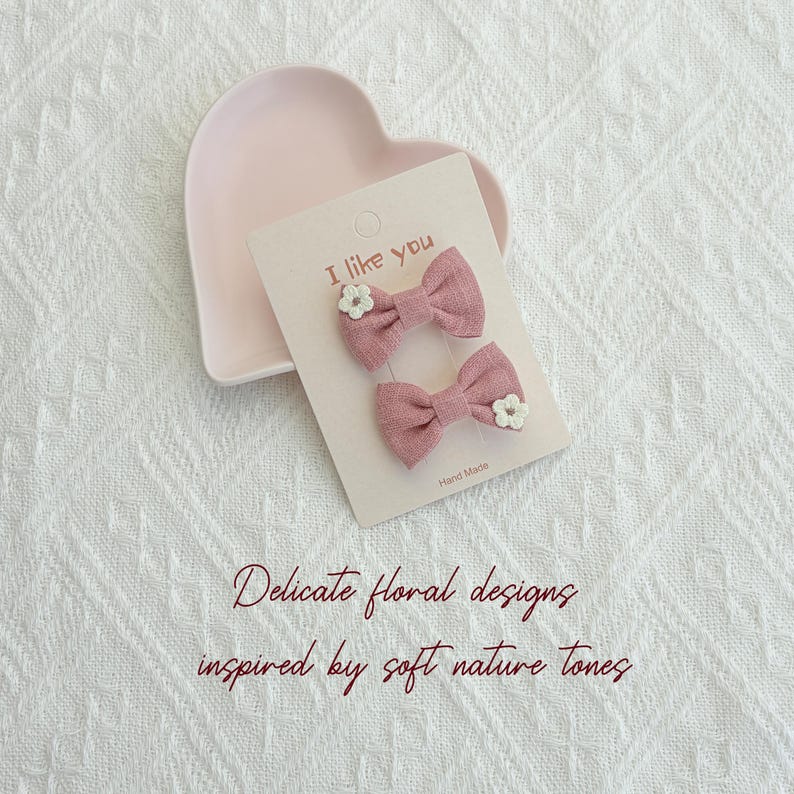 May include: Two pink bow hair clips with white floral accents are displayed on a light pink card that reads "I like you". The card is placed on a heart-shaped dish. The text "Delicate floral designs inspired by soft nature tones" is also visible.