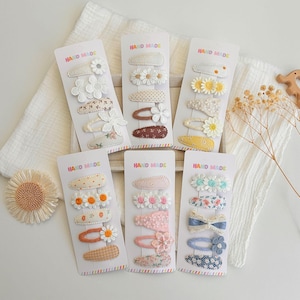 May include: Six white cardboard display cards labeled HAND MADE, each holding five hair clips. The clips feature various designs including embroidered daisies, floral patterns, polka dots, and solid colors in shades of white, yellow, pink, and blue.