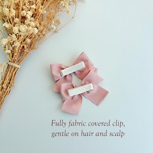 Peut inclure: Deux n&oelig;uds pour cheveux en tissu rose poudr&eacute; avec des pinces enti&egrave;rement recouvertes de tissu au dos. Le texte indique : Fully fabric covered clip, gentle on hair and scalp. Les n&oelig;uds ont une finition textur&eacute;e et sont pr&egrave;s de fleurs s&eacute;ch&eacute;es.
