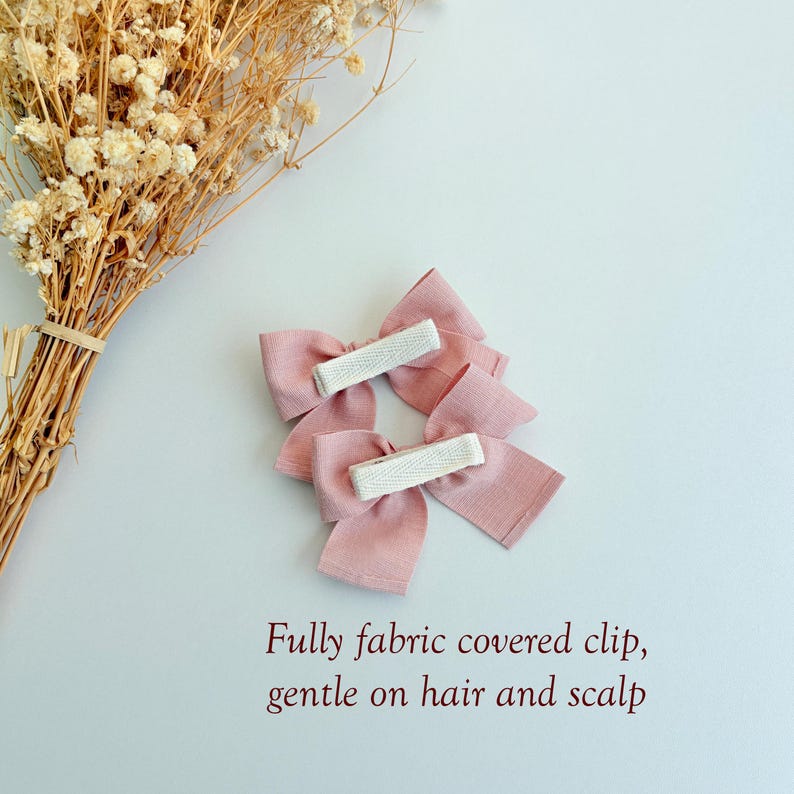 Peut inclure: Deux n&oelig;uds pour cheveux en tissu rose poudr&eacute; avec des pinces enti&egrave;rement recouvertes de tissu au dos. Le texte indique : Fully fabric covered clip, gentle on hair and scalp. Les n&oelig;uds ont une finition textur&eacute;e et sont pr&egrave;s de fleurs s&eacute;ch&eacute;es.