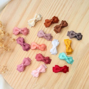 May include: A collection of small hair clips featuring bows made from ribbed grosgrain ribbon in various colors including cream, brown, pink, peach, white, yellow, teal, and red, each attached to a metal alligator clip base for secure attachment.