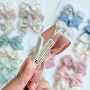 Peut inclure: Un n&oelig;ud pour cheveux en tissu rose poudr&eacute; fix&eacute; sur une pince crocodile en m&eacute;tal blanc. Le n&oelig;ud pr&eacute;sente une mati&egrave;re tiss&eacute;e textur&eacute;e et est pr&eacute;sent&eacute; avec une collection d'autres n&oelig;uds pour cheveux en tissu de couleurs et motifs vari&eacute;s.