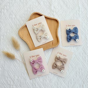 May include: Four pairs of handmade hair bows in various colors: beige, blue, mauve, and taupe. Each bow is adorned with a small white flower and attached to a card that reads "I like you".