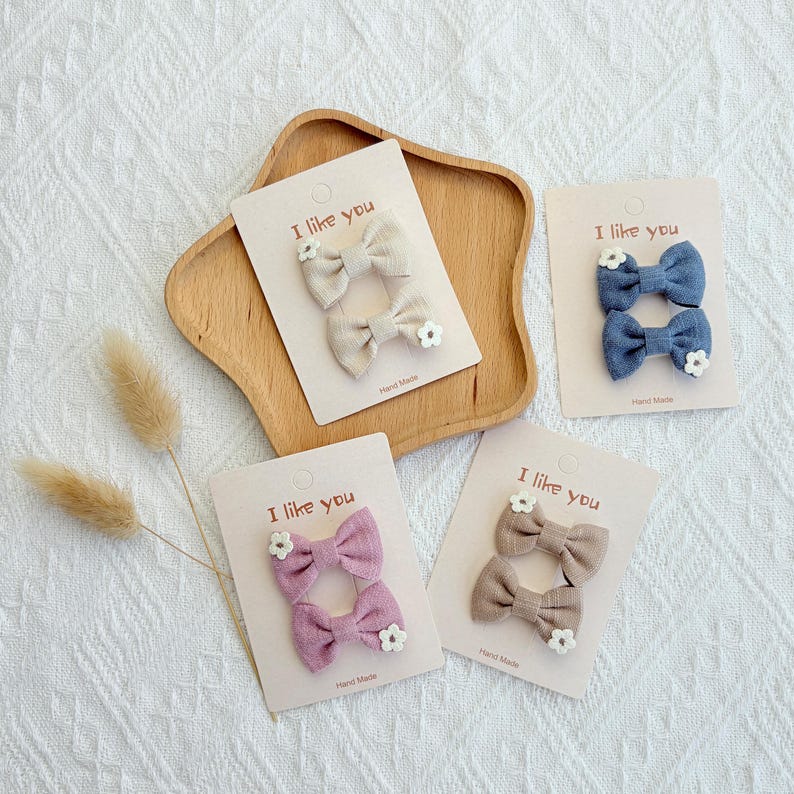 May include: Four pairs of handmade hair bows in various colors: beige, blue, mauve, and taupe. Each bow is adorned with a small white flower and attached to a card that reads "I like you".