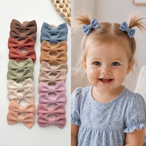 May include: A collection of small fabric hair bows in various solid colours including brown, blue, tan, green, cream, and pink, each attached to a thin elastic band. The bows feature a textured fabric finish and are arranged in two vertical columns.