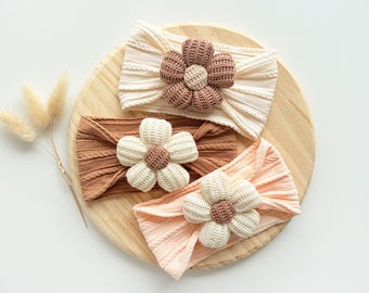 May include: Three knit headbands in cream, brown, and light peach, each featuring a large, dimensional flower applique. The cream headband has a brown flower center, the brown headband has a cream center, and the peach headband has a brown center.