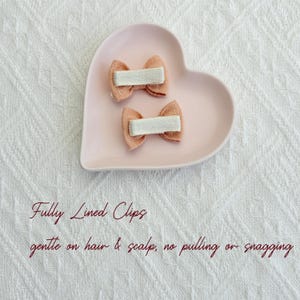May include: Two small, peach-colored hair bows with white ribbon centers rest on a pink heart-shaped dish. The bows are fully lined and gentle on hair and scalp, with no pulling or snagging.