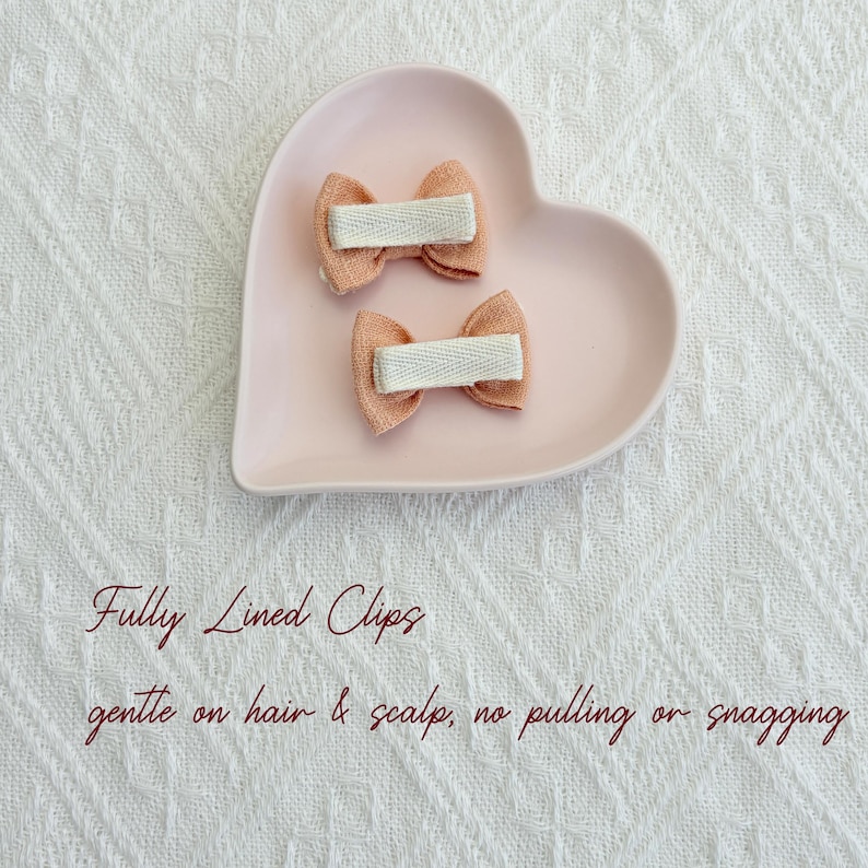 May include: Two small, peach-colored hair bows with white ribbon centers rest on a pink heart-shaped dish. The bows are fully lined and gentle on hair and scalp, with no pulling or snagging.