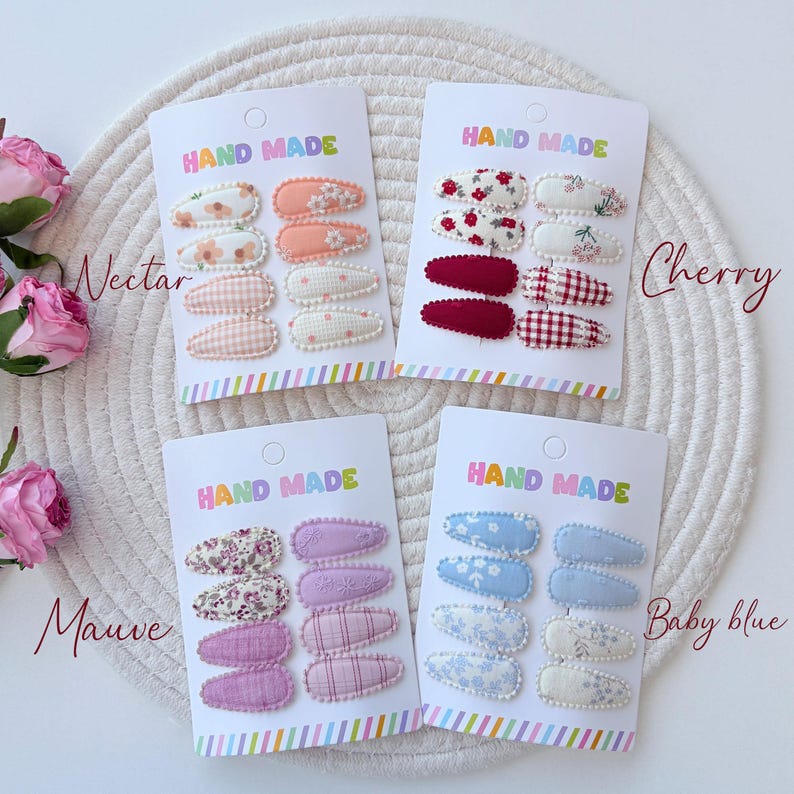 Op de afbeelding: Vier sets van acht met stof beklede haarclips op witte kaarten met het opschrift HAND MADE. De sets bevatten diverse patronen, waaronder bloemen, ruitjes en effen kleuren in de kleurenpaletten nectar, kers, mauve en babyblauw.