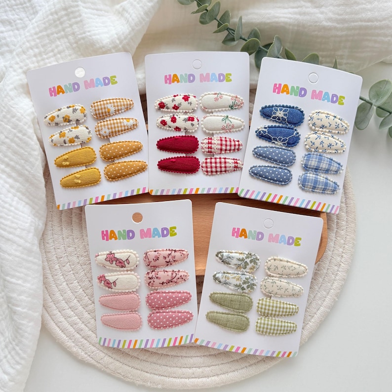 Op de afbeelding: Vijf witte presentatiekaarten met het label HAND MADE, elk met acht met stof beklede haarspeldjes. De speldjes hebben diverse patronen, waaronder bloemen, ruitjes, stippen en effen kleuren in gele, rode, blauwe, roze en groene paletten.