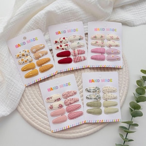 Op de afbeelding: Vijf witte presentatiekaarten met het opschrift HAND MADE, elk met acht met stof beklede haarspeldjes. De speldjes hebben diverse patronen, waaronder bloemenprints, vichy-ruitjes en effen kleuren in tinten geel, rood, roze, groen en cr&egrave;me.
