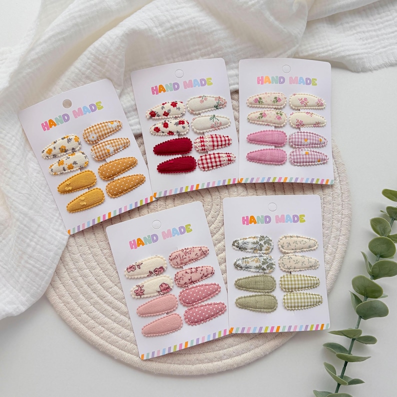 Op de afbeelding: Vijf witte presentatiekaarten met het opschrift HAND MADE, elk met acht met stof beklede haarspeldjes. De speldjes hebben diverse patronen, waaronder bloemenprints, vichy-ruitjes en effen kleuren in tinten geel, rood, roze, groen en cr&egrave;me.