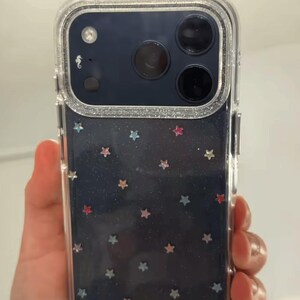 May include: A clear smartphone case with a silver glitter border and a dark blue background featuring scattered small multi-colored star shapes. The case includes a cutout for a triple-lens camera system and a small white seahorse icon.