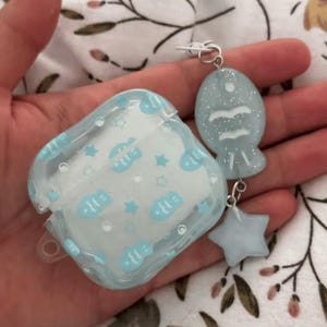 May include: A clear protective case for wireless earbuds featuring a light blue pattern of small fish and stars. Attached to the side is a silver-toned metal ring holding a light blue glittery fish-shaped charm and a small light blue star charm.