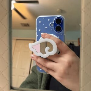May include: A blue smartphone case featuring a white star pattern and a white, wing-shaped phone grip attachment. The case has a clear border and a circular cutout for the dual camera lenses, providing a protective cover for the mobile device.