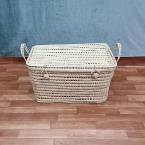 Può includere: Un cesto rettangolare intrecciato con coperchio e manici. Il cesto è realizzato in materiale intrecciato di colore naturale. Il coperchio ha un motivo intrecciato e le maniglie sono ad anello. Il cesto è appoggiato su una superficie di legno.