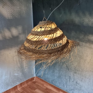 Puede incluir: Una lámpara colgante cónica artesanal hecha de fibras naturales tejidas. La lámpara tiene un diseño en espiral y un fondo con flecos. La luz está suspendida de un cable blanco e ilumina el interior, creando un brillo cálido.