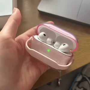 May include: A pair of white wireless earbuds housed in a translucent pink protective charging case. The case is open, revealing the earbuds inside, and features a small, illuminated green LED indicator light on the front exterior surface.