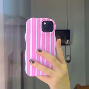 May include: A mobile phone case featuring a wavy, irregular edge design with vertical pink and white stripes. The case has a cutout for a dual-lens camera system on the back, showcasing a glossy finish and a distinct, non-uniform, undulating shape.