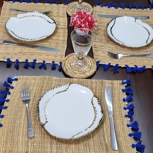 Puede incluir: Una mesa puesta con platos blancos con ribete dorado, cubiertos de acero inoxidable y manteles individuales tejidos con ribete de borlas azules. También hay un vaso transparente con borde dorado y un pequeño arreglo floral rojo.