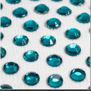 May include: Close-up of teal blue rhinestones on a white fabric background. The rhinestones are arranged in a grid pattern.