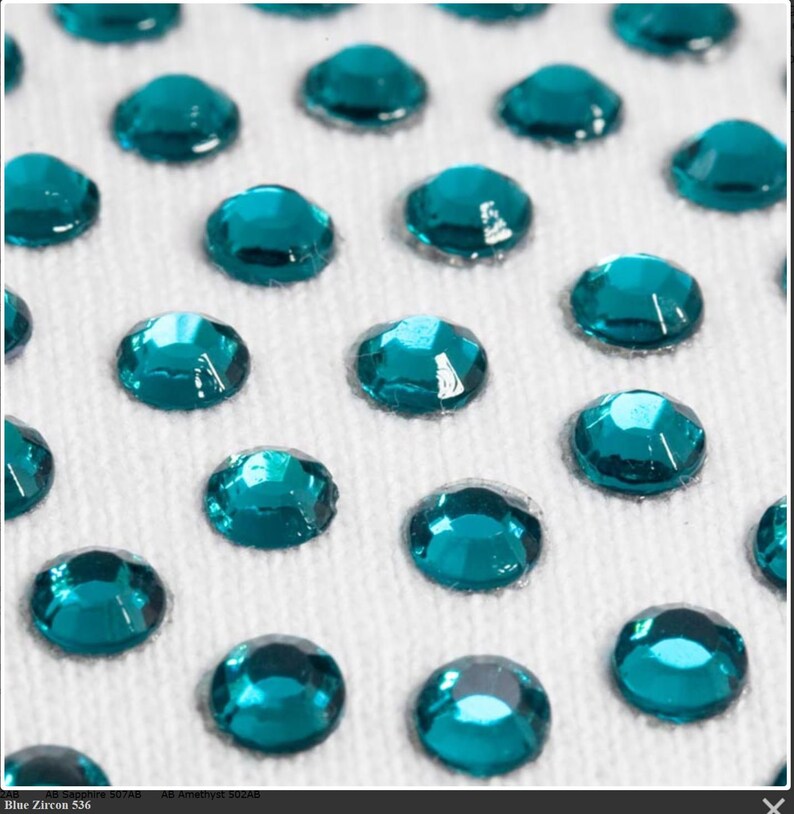 May include: Close-up of teal blue rhinestones on a white fabric background. The rhinestones are arranged in a grid pattern.
