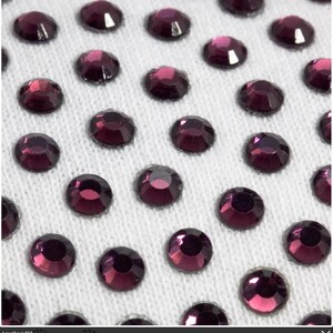 May include: Close-up of a white fabric surface covered in small, round, purple rhinestones.