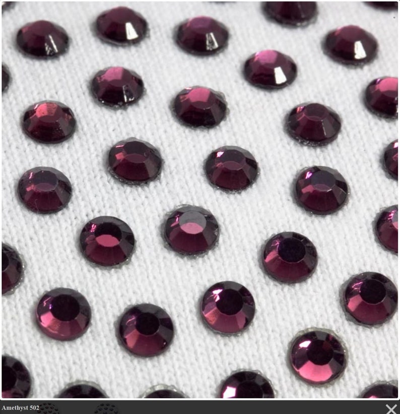May include: Close-up of a white fabric surface covered in small, round, purple rhinestones.