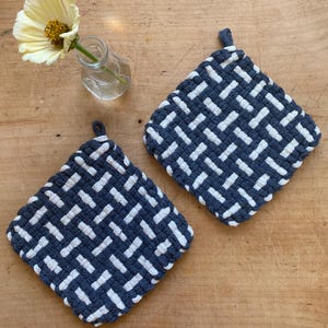 May include: Two navy blue and white woven potholders with hanging loops. A small glass vase holds a yellow flower. The potholders feature a geometric pattern and are set on a wooden surface. The potholders are approximately 20 cm square.
