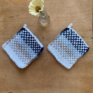 May include: Two square woven potholders with a loop for hanging. The potholders feature a woven pattern in white, navy blue, and tan. A small glass vase with a yellow flower sits nearby on a wooden surface.