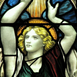 Puede incluir: Una ventana de vitral que representa a un ángel con los brazos extendidos, vestido con una túnica verde y un halo de color amarillo y naranja. El ángel tiene el pelo largo y rubio y una expresión serena.