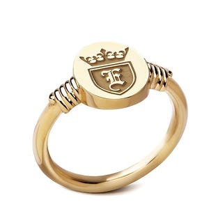 Peut inclure: Bague dorée avec une face ovale ornée d'une couronne et de la lettre "E" dans un écusson. L'anneau de la bague est lisse, et la face est reliée à l'anneau par un motif enroulé. Un bijou classique.