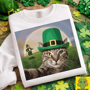 Pode incluir: Camisola branca com um design do Dia de São Patrício. Apresenta um gato usando um chapéu verde, tirando uma selfie. O design inclui um duende e um arco-íris. O design é cercado por trevos e moedas de ouro.