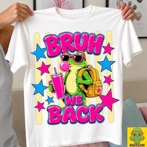 May include: A white t-shirt featuring a graphic of a green frog wearing pink sunglasses, blowing a pink bubble, and carrying a yellow backpack. The frog holds a pink tumbler. The text reads BRUH WE BACK in bold pink letters, surrounded by blue and pink stars.