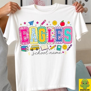 May include: A white t-shirt featuring the word EAGLES in colorful, patterned block letters. The design includes school-themed graphics like a yellow bus, books, a pencil, an apple, and a smiley face, with the text school name written below.