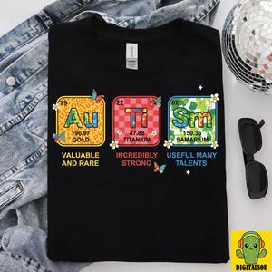 May include: Black t-shirt featuring colorful periodic table elements: Gold (Au), Titanium (Ti), and Samarium (Sm). Each element is in a square with its atomic number, symbol, and description. Butterflies and flowers decorate the design. The text reads "VALUABLE AND RARE", "INCREDIBLY STRONG", and "USEFUL MANY TALENTS".
